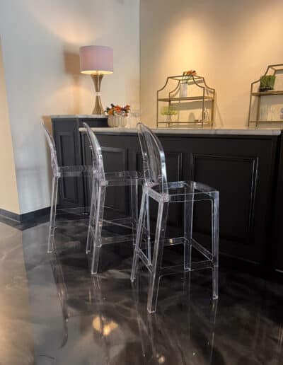 Two transparent acrylic bar stools in front of a dark bar, with a pink lamp and decorative glass shelves on the countertop.