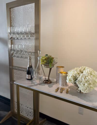 A display with a decorative screen holding glassware, adjacent to a small table with a champagne bottle and hydrangeas.