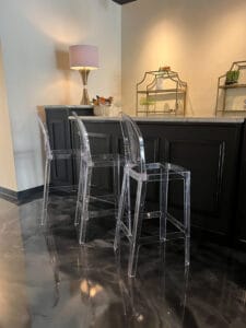Two transparent acrylic bar stools in front of a dark bar, with a pink lamp and decorative glass shelves on the countertop.