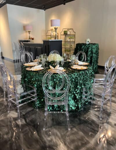 Elegant tables covered in green tablecloths, with clear acrylic chairs, in a modern room with grey floors.