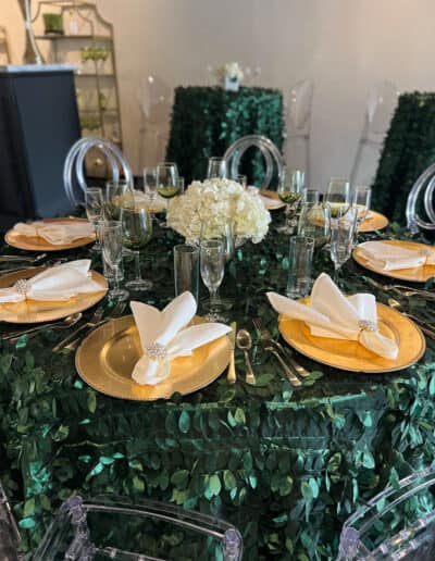Elegant dining table with gold-rimmed plates, crystal glassware, and a white hydrangea centerpiece on a leafy green tablecloth.