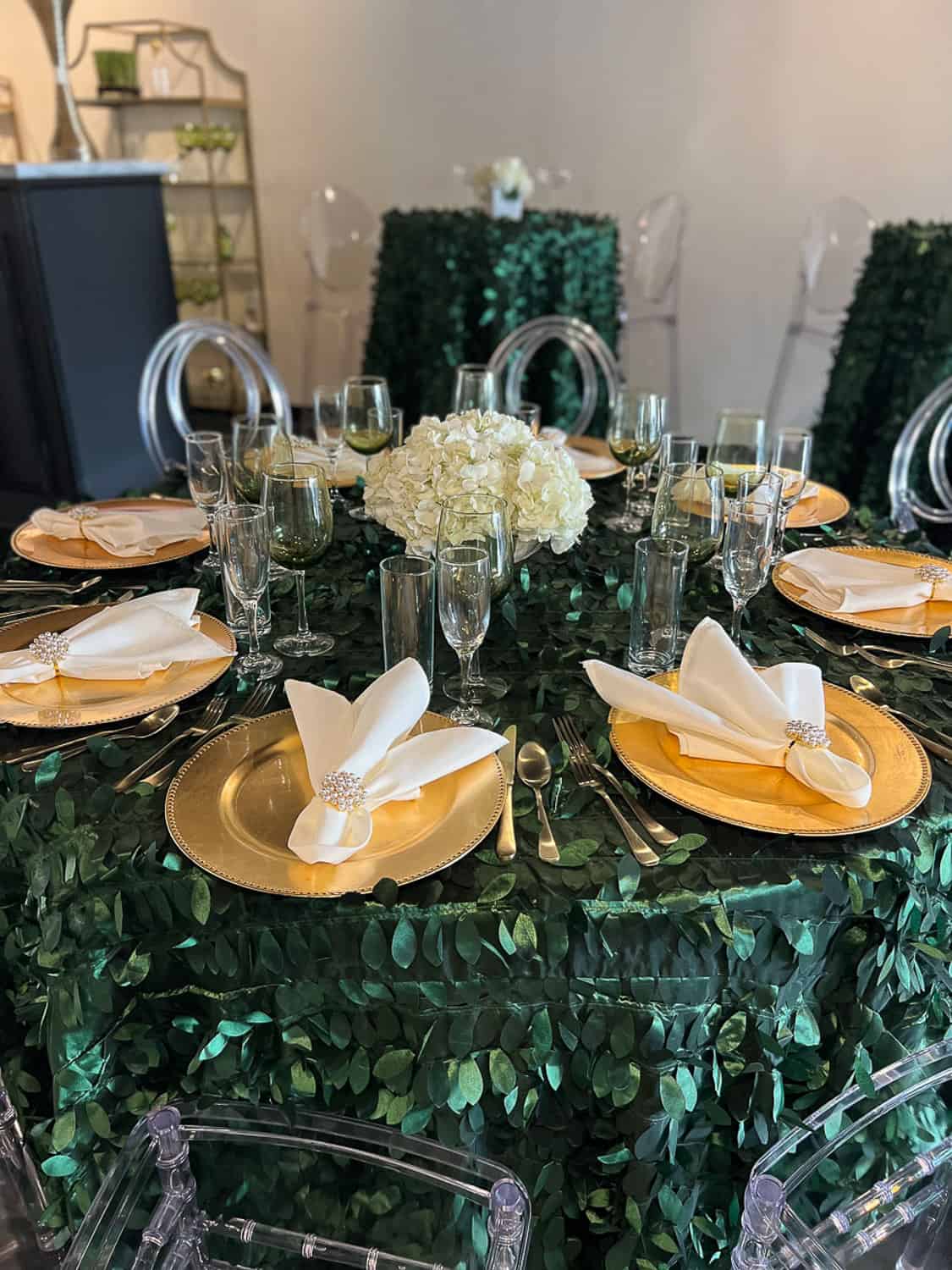 Elegant dining table with gold-rimmed plates, crystal glassware, and a white hydrangea centerpiece on a leafy green tablecloth.