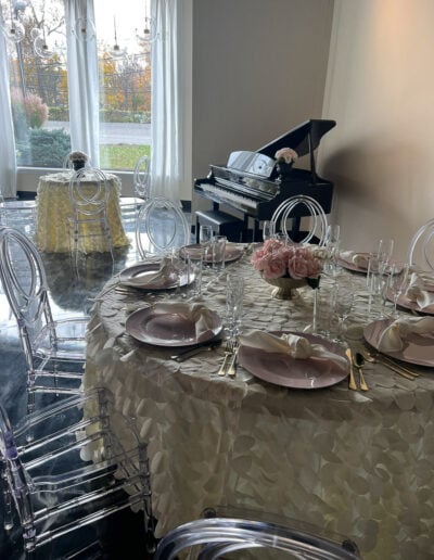 A round table covered in a white tablecloth, a vase with pink flowers, transparent chairs, and a piano.