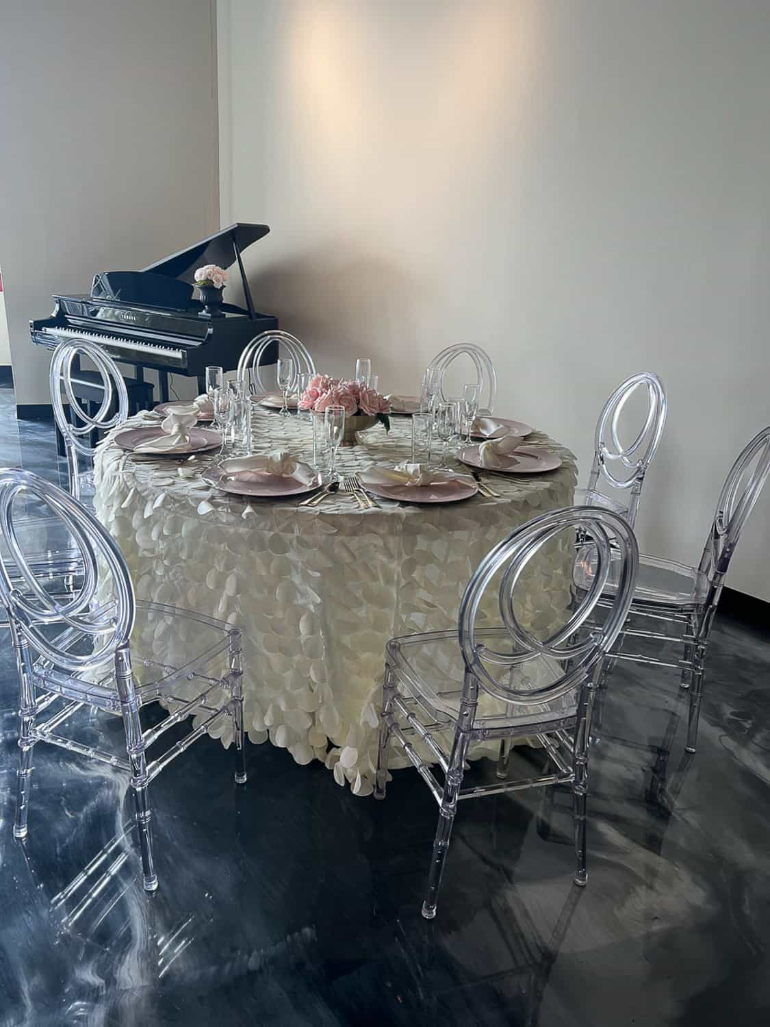 A round table covered in a white tablecloth, a floral centerpiece, transparent chairs, and a piano.