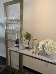 A display with a decorative screen holding glassware, adjacent to a small table with a champagne bottle and hydrangeas.
