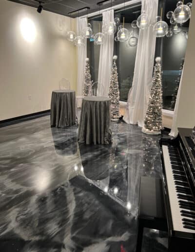 Event space with black floor, decorated with white Christmas trees, round tables, hanging lights; and a piano to the side.
