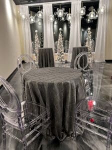 An elegant event space with a small round table, gray tablecloth, acrylic chairs, surrounded by mirrors and decorative lights.