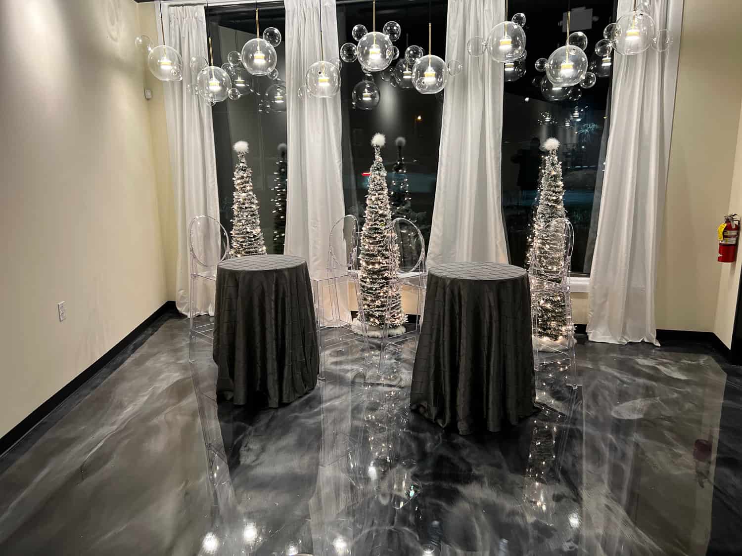 Modern event space with sheer curtains, shiny floor, and tables with metallic spiral centerpieces under hanging circular lights.