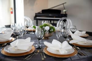 Dining table set with white and gold plates, wine glasses, and a centerpiece, with a piano in the corner.