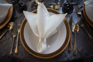 Table setting with a folded white napkin, on a golden-rimmed white plate, surrounded by gold cutlery on a dark tablecloth.