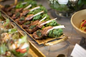 Plated lamb chops garnished with lettuce, presented in a buffet setup with various food in the background.