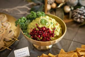 A bowl of pomegranate guacamole garnished with lime, surrounded by chips and decorative items on a festive table.