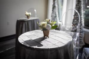 A round table with a gray tablecloth and a small floral centerpiece, accompanied by decorative trees in the background.