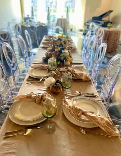 Elegantly set dining table with clear chairs and golden tableware, decorated with pumpkins and greenery.