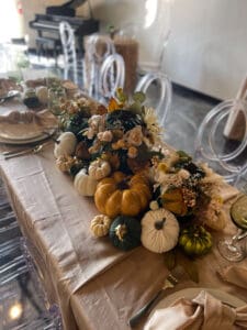 Elegant dining table set with a centerpiece with assorted decorative pumpkins and flowers, surrounded by clear acrylic chairs.