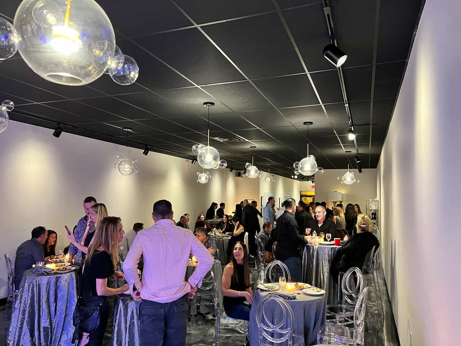 People mingling and dining in a modern event space with round tables, clear chairs, and spherical hanging lights.