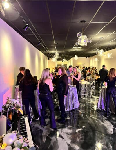 Guests socialize in a well-lit event space with modern décor and marble floors, with round tables and overhead spherical lights.