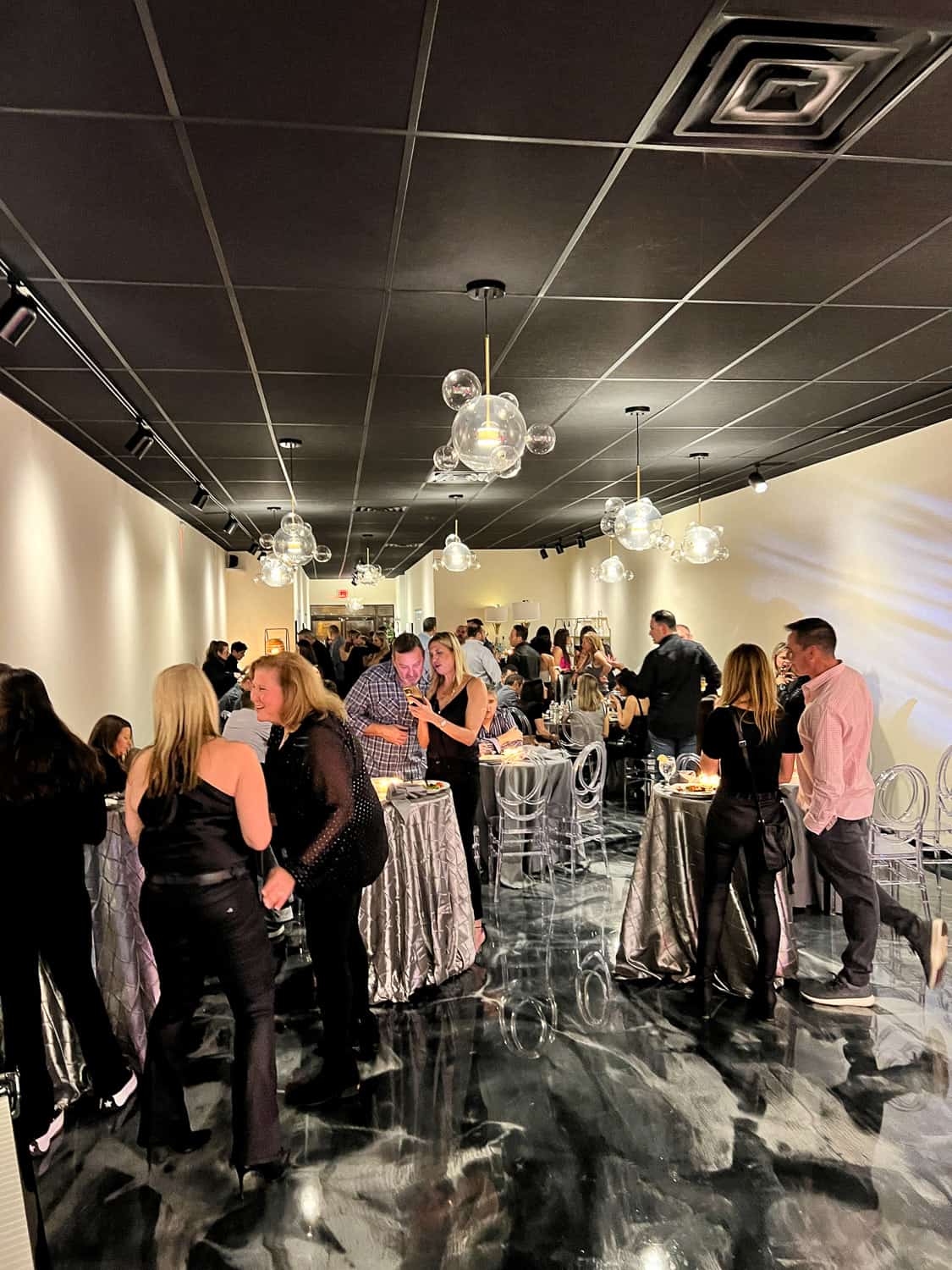 People socializing at a formal indoor event with elegant decor, featuring marble floors and modern hanging lights.