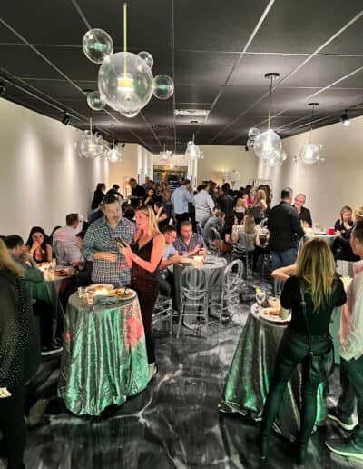 Crowded indoor event with people socializing around tables, elegant lighting, and a modern décor.