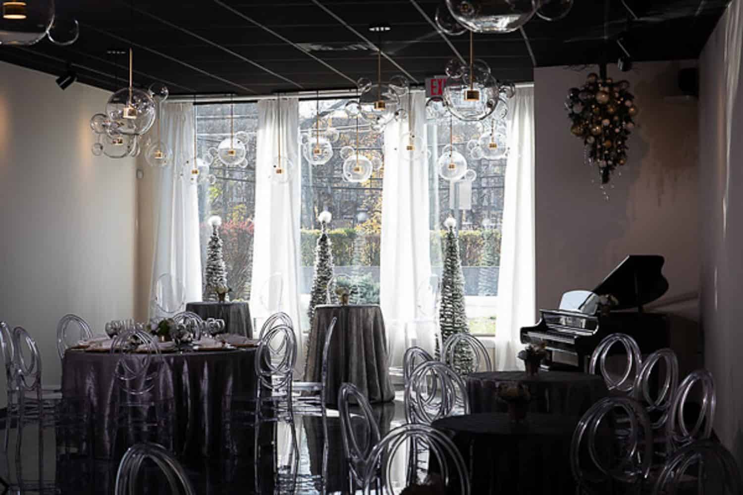 Elegant dining room with circular chairs, decorated tables, a piano, and large windows with a view of greenery outside.