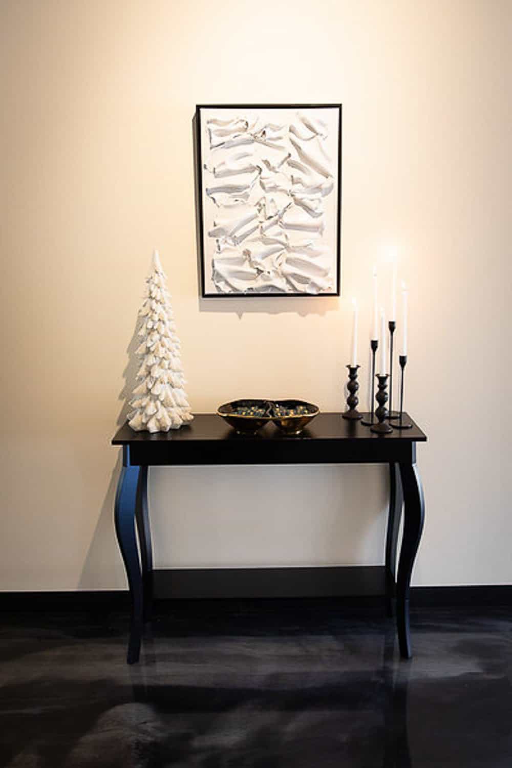 Decor featuring a black table set with a white ceramic Christmas tree, candlesticks, and a bowl, in front of abstract wall art.