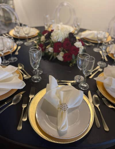 Table setting with a black tablecloth, gold-rimmed plates, red and white floral centerpiece, gold flatware and crystal glassware.