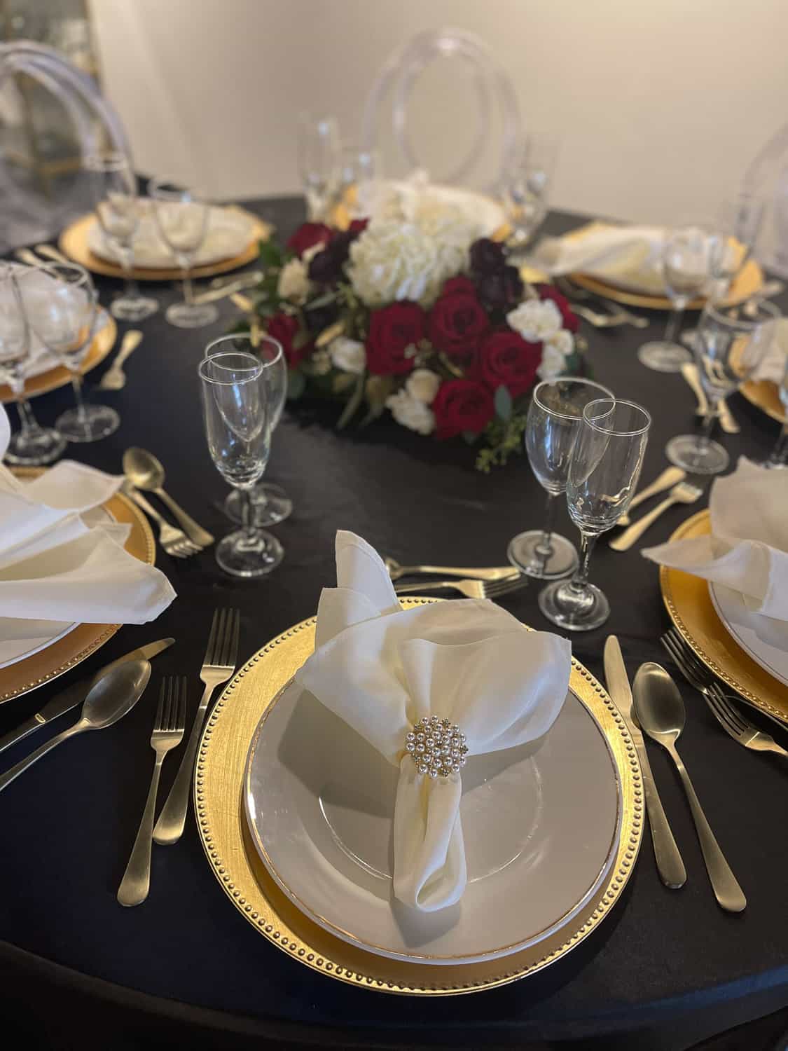 Table setting with a black tablecloth, gold-rimmed plates, red and white floral centerpiece, gold flatware and crystal glassware.