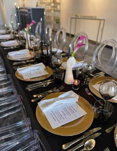 Elegant dining table setup with black tablecloth, golden chargers, clear stemware, and pink cala lily accents.