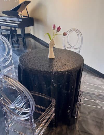 A small round table covered in a black tablecloth, a vase holding a single tulip, three transparent chairs, and a piano.