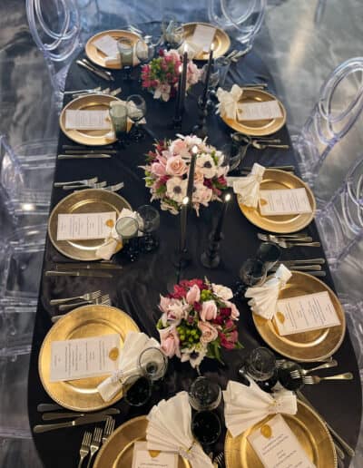 Elegant dining table set for ten with gold plates, crystal glassware, and floral centerpieces on a black tablecloth.