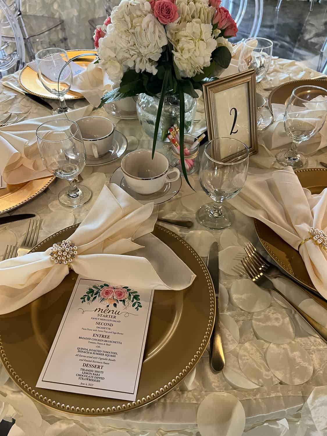 Dining table setting for a formal event with floral centerpiece, gold-rimmed plates, menu cards, and numbered table sign.