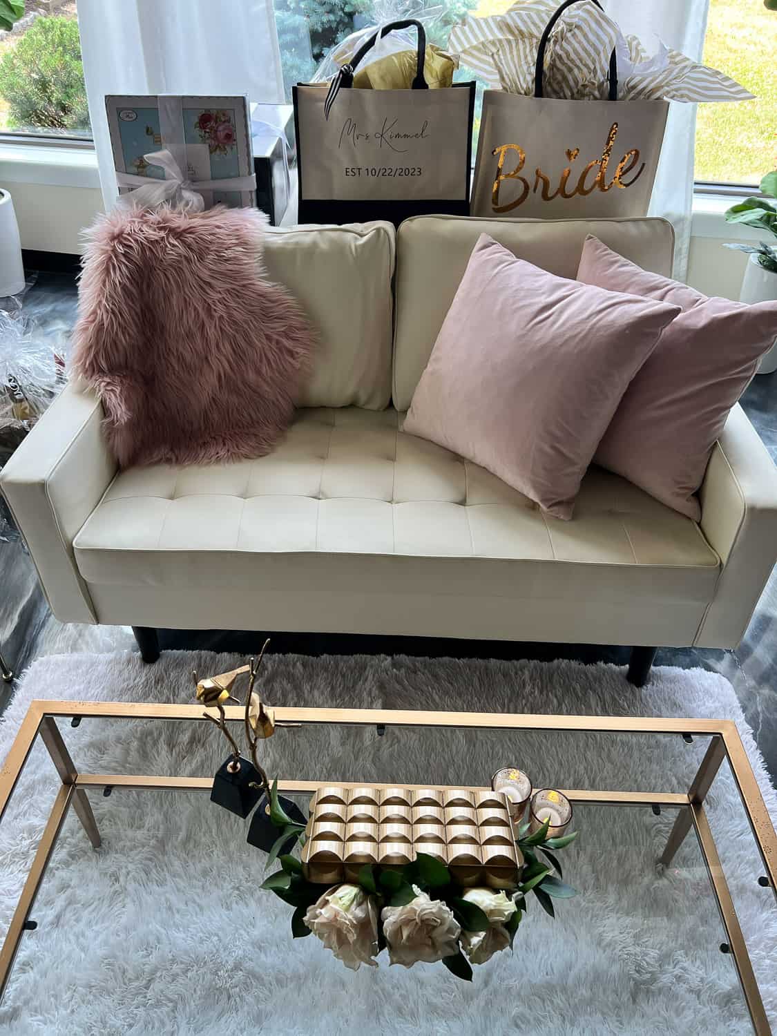 Cozy living room-like corner with a white sofa and pink pillows, a glass coffee table, a fur rug, and various gift bags.