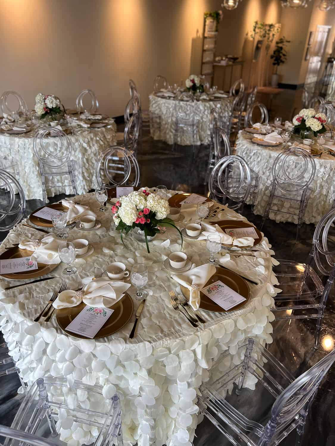 Elegant wedding reception setup with round tables, floral centerpieces, transparent chairs, and place settings.