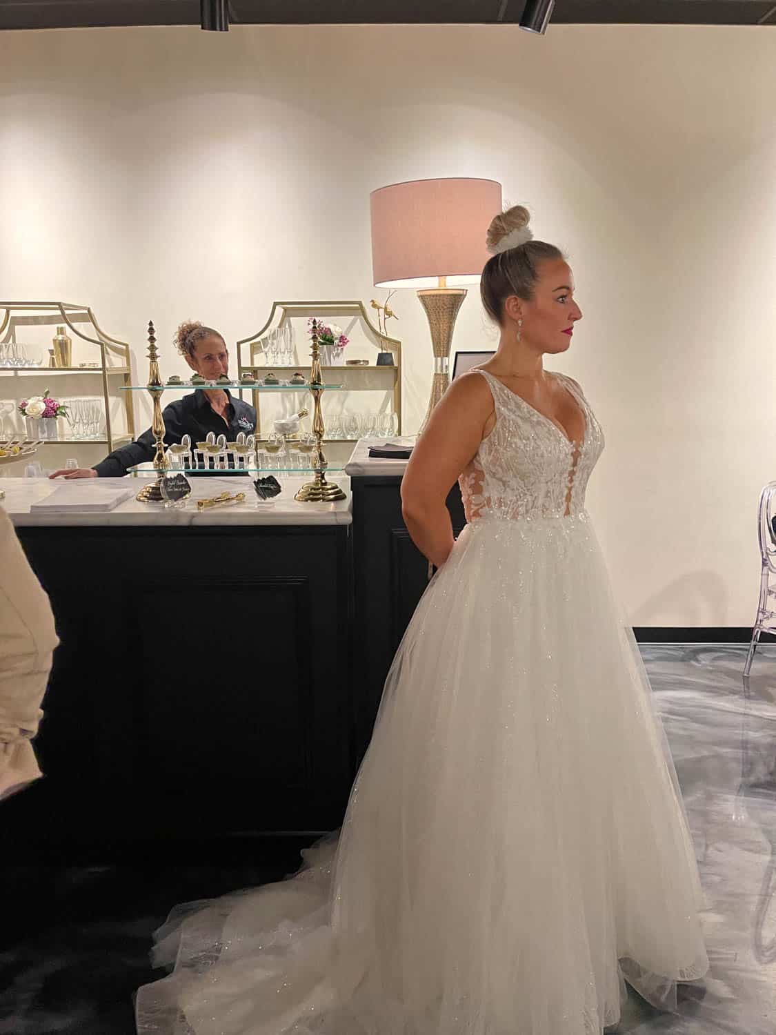 A woman in a white wedding dress stands at a reception with elegant decor, mirrored surfaces, and another woman at the bar.