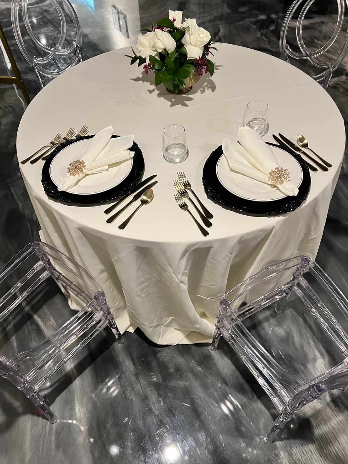 Round dining table set for two with white and black plates, silver cutlery, clear chairs, and a centerpiece of white flowers.