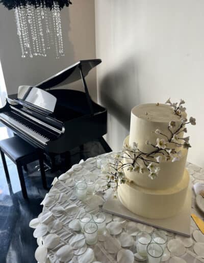A tiered white cake with floral decorations on a table, surrounded by candles, with a grand piano in the background.