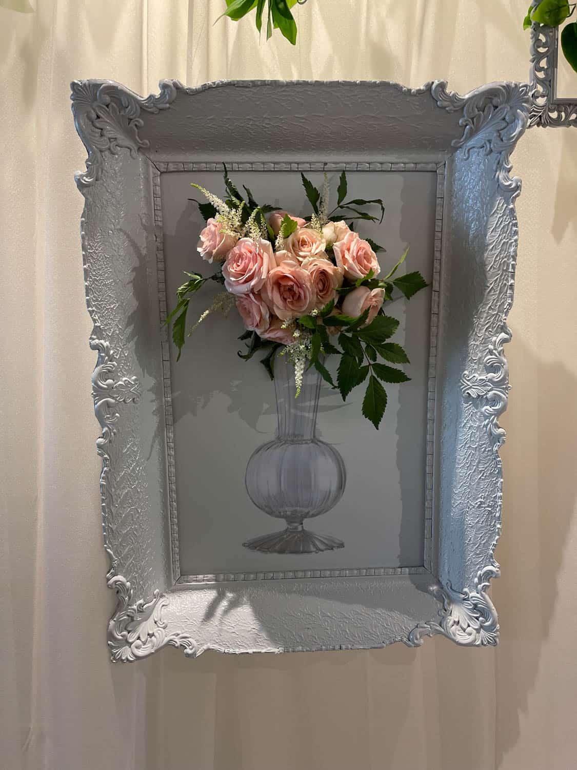 An ornate silver frame displaying a three-dimensional artistic arrangement of pink roses in a vase, set against a pale wall.