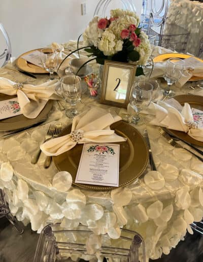 Dining table setting for a formal event with floral centerpiece, gold-rimmed plates, menu cards, and numbered table sign.