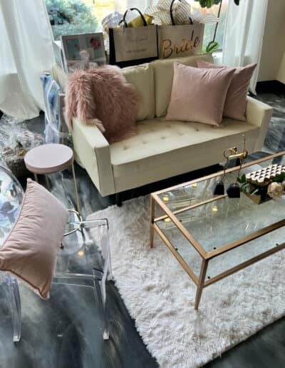 Cozy living room-like corner with a white sofa and pink pillows, a glass coffee table, a fur rug, and various gift bags.