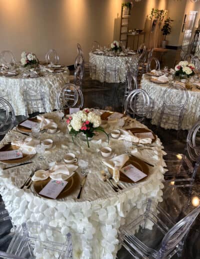 Wedding reception with round tables covered in white tablecloths, clear chairs, and centerpieces of white and red flowers.