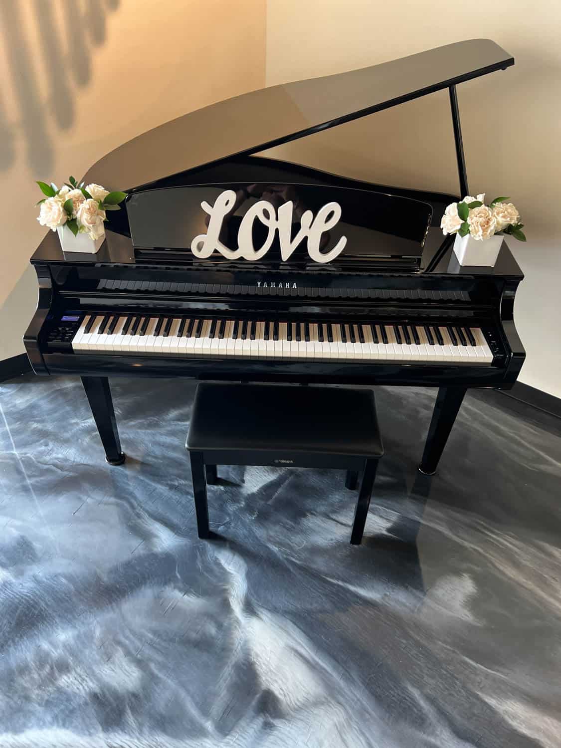 Grand piano with the word "love" displayed on music stand, flanked by floral arrangements, sitting on a marbled grey floor.