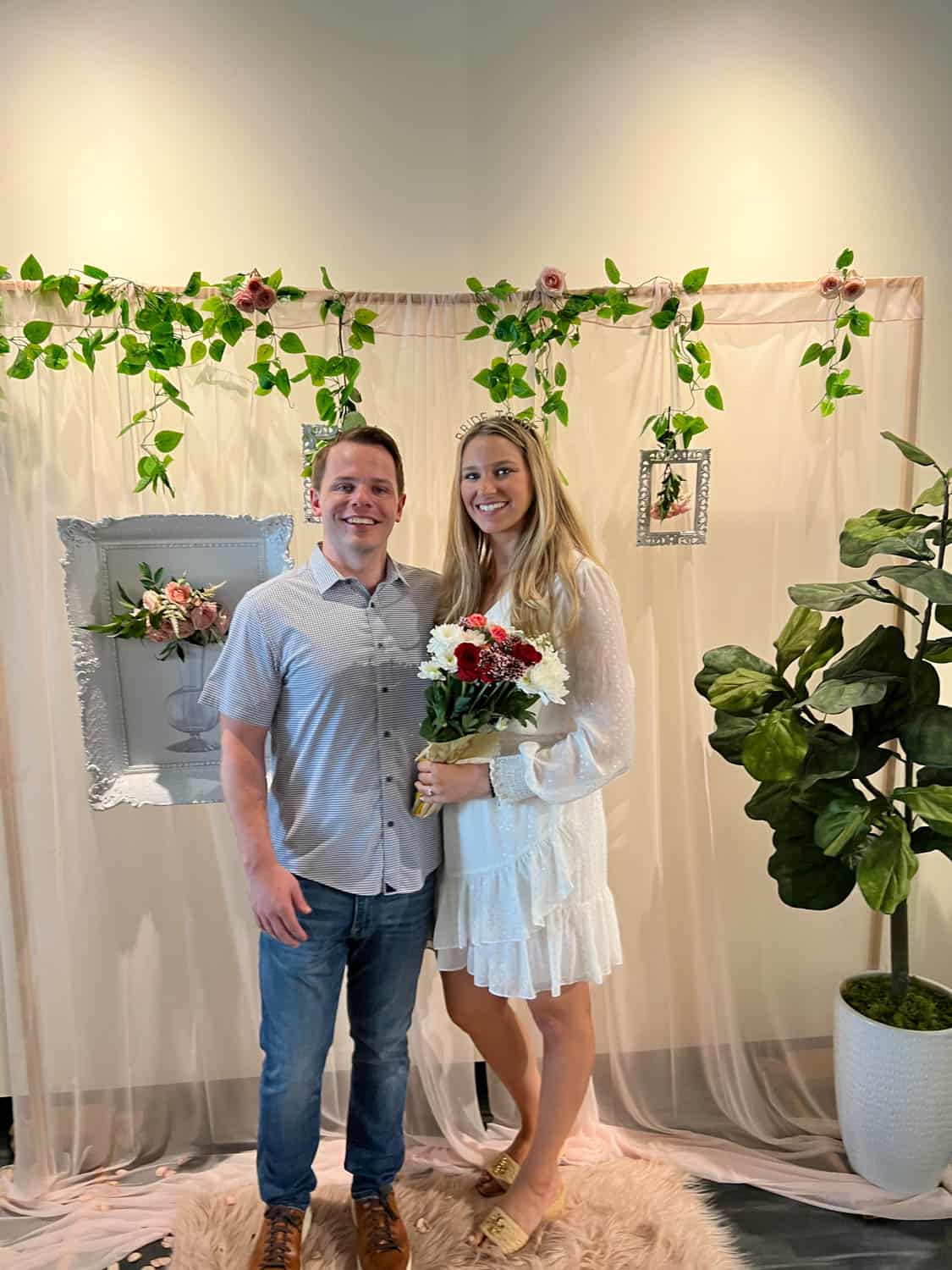 A couple posing with flowers in a decorated indoor space with a framed backdrop, plant, and drapery.