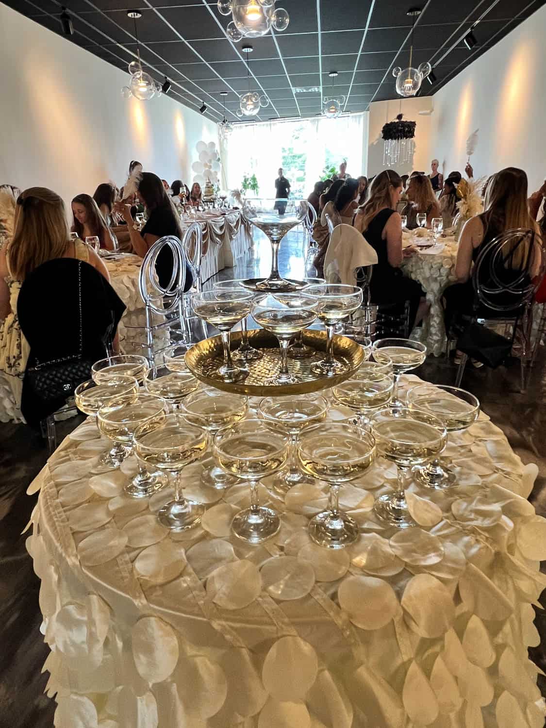 Table with champagne glasses set in the foreground at a wedding reception venue, with guests seated at tables in background.
