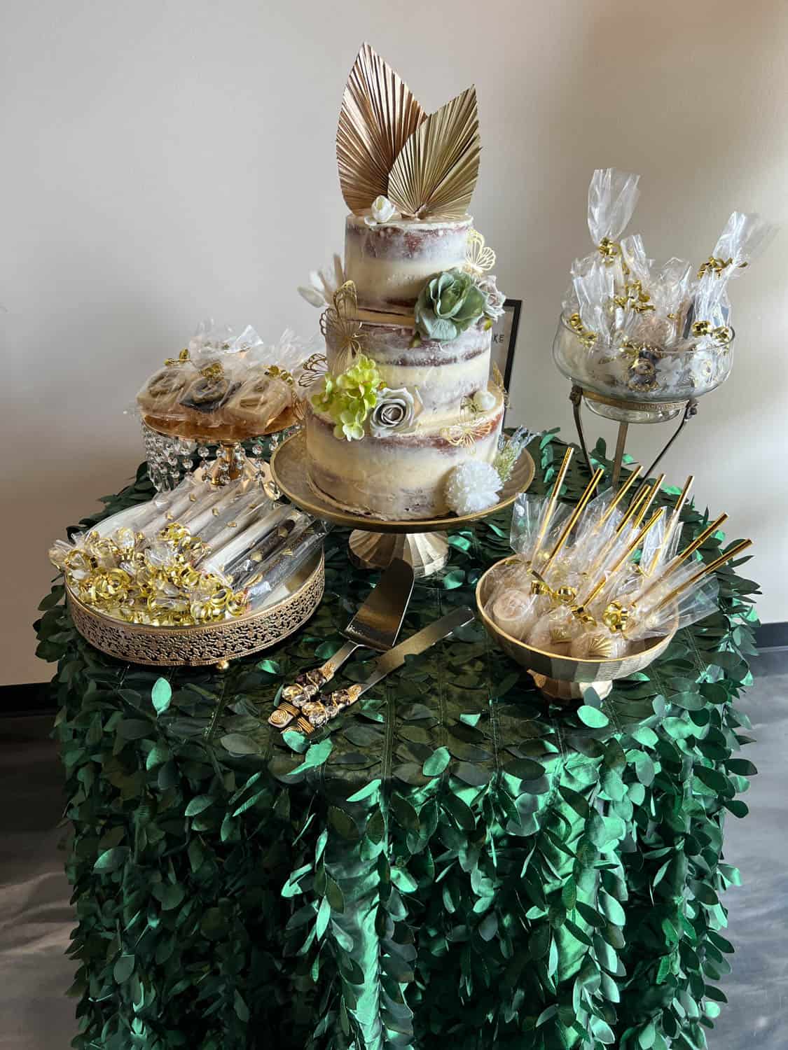 A dessert table featuring a tiered cake with gold leaves and flowers, surrounded by various trays of decorated confections.