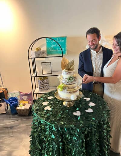 A couple cutting a three-tier cake at a reception, with decorations and a congratulatory sign with their names and date.