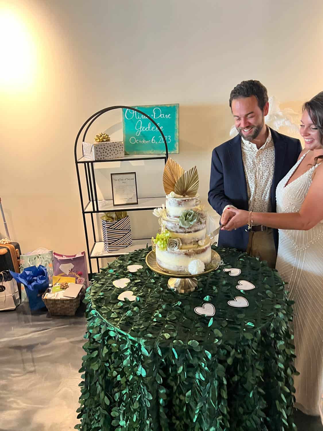 A couple cutting a three-tier cake at a reception, with decorations and a congratulatory sign with their names and date.